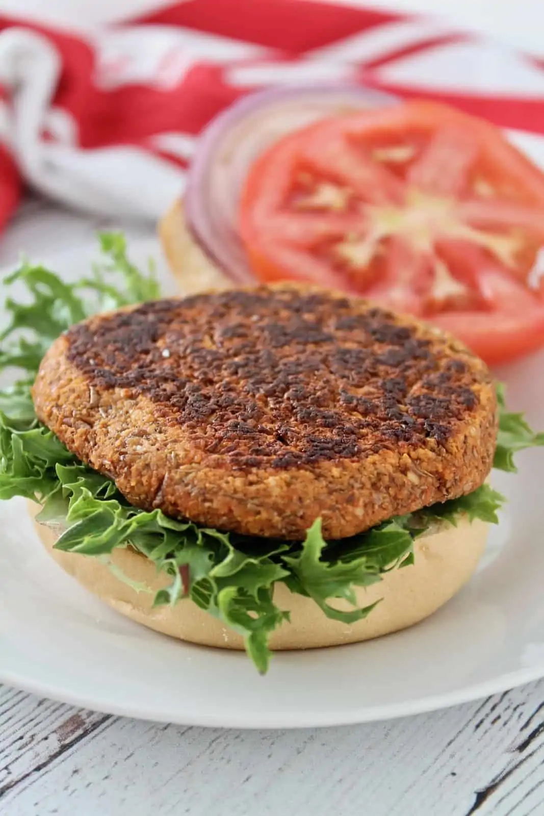 Close up of open faced sprouted lentil burger on green lettuce on a bun on a white plate with tomato and onion in the background
