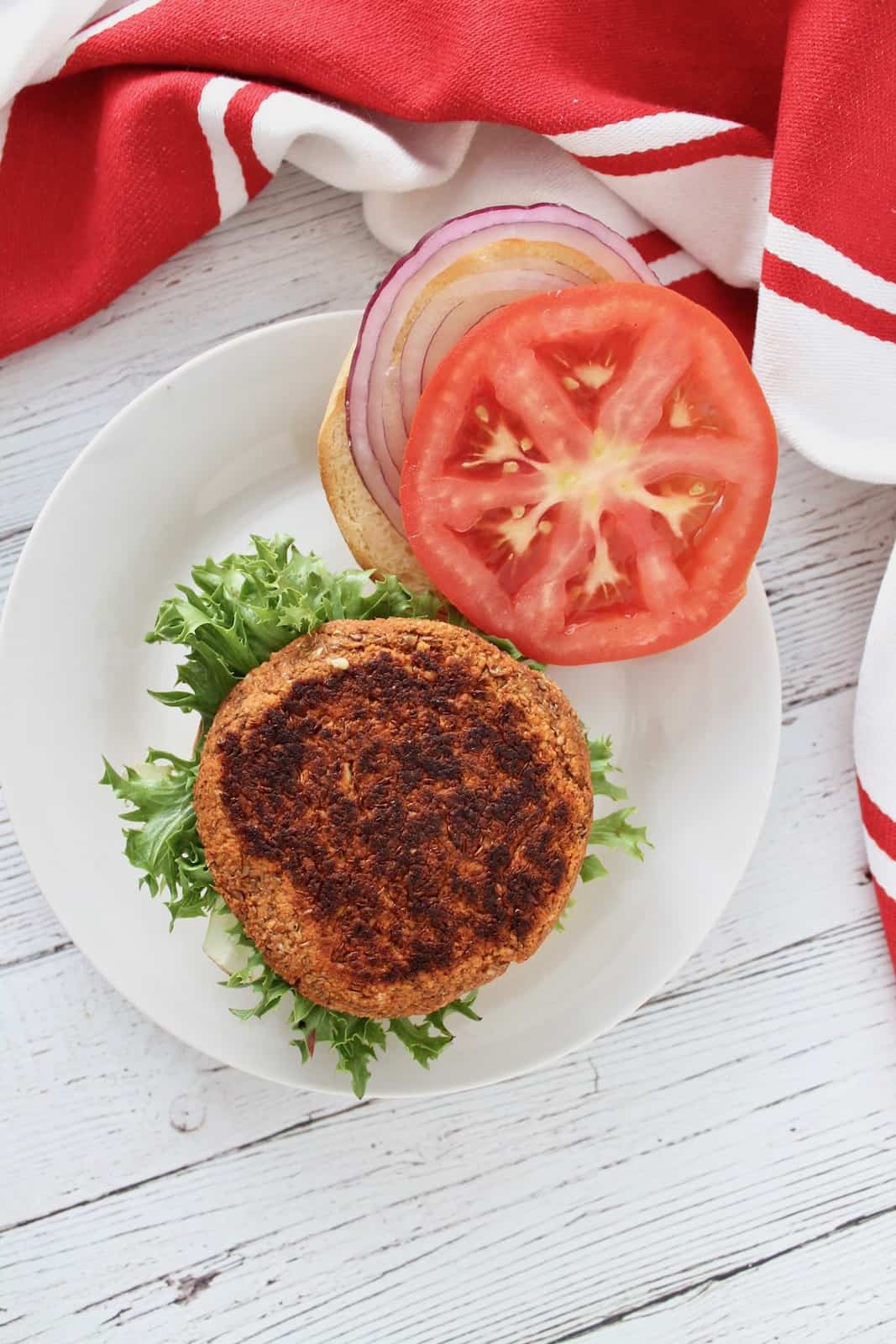 Overhead shot of open faced sprouted lentil burger on green lettuce on a bun on a white plate with tomato and onion next to it on a white plate next to a red and white striped dish towel