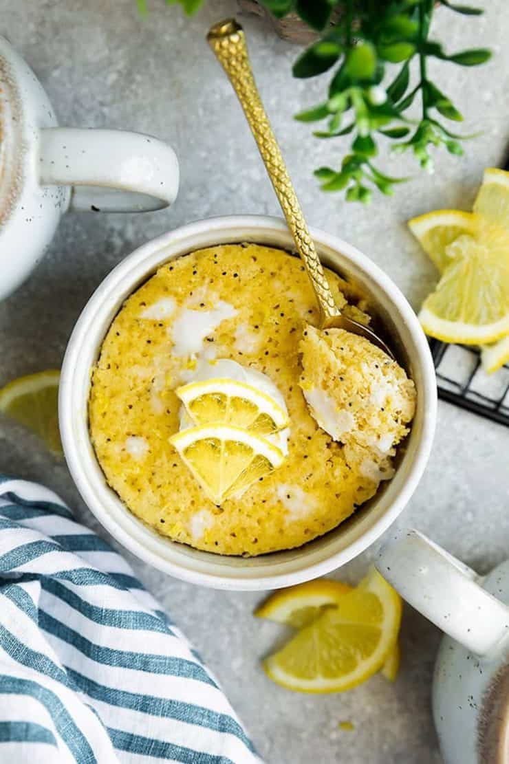 Overhead shot of white mug filled with yellow lemon poppyseed cake with lemon slices and white frosting on top with a gold spoon in it
