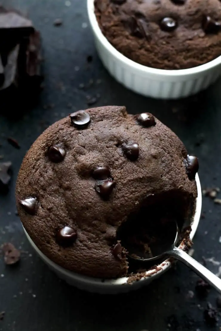 Overhead shot of white ramekin with chocolate cake inside with chocolate chips on top on a dark grey surface