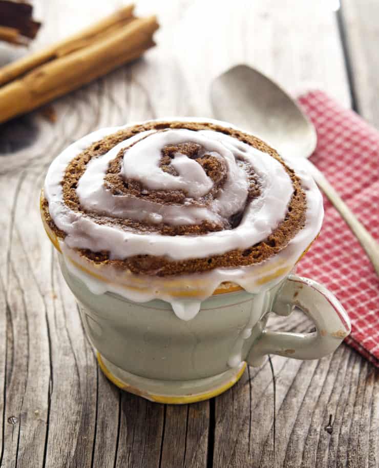 Mug filled with cinnamon cake with white frosting in a swirl design on top on a wooden table next to a red and white checkered dish towel with a spoon on it next to cinnamon sticks