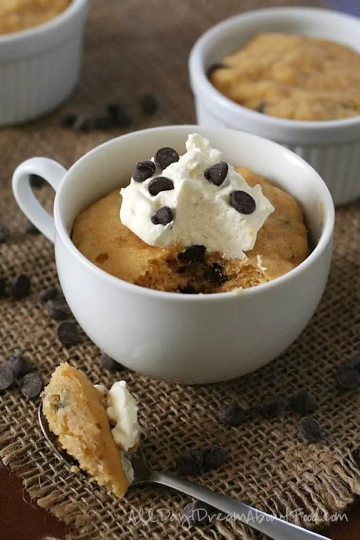 White mug filled with peanut butter cake topped with whipped cream and chocolate chips on top of burlap sprinkled with chocolate chips next to a spoon with a bite of cake on it