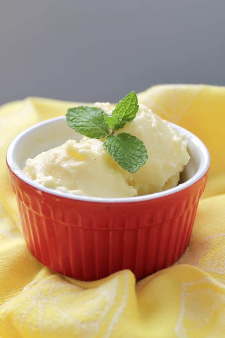 Red ramekin with yellow pineapple sorbet in it with a mint leaf on top sitting on a yellow dish towel