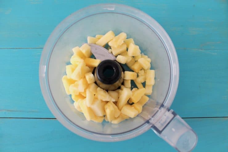 Overhead shot of small pieces of pineapple in a food processor on a blue wooden surface