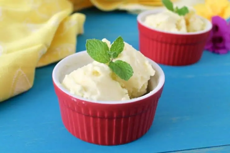 Small red ramekin with pineapple sorbet in it with a mint leaf on top with another ramekin in the background next to a yellow dish towel on a blue wooden surface