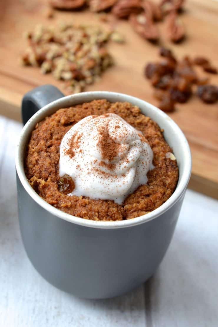 Grey mug filled with carrot mug cake with a dollop of white whipped cream on top next to a wooden cutting board with nuts on top