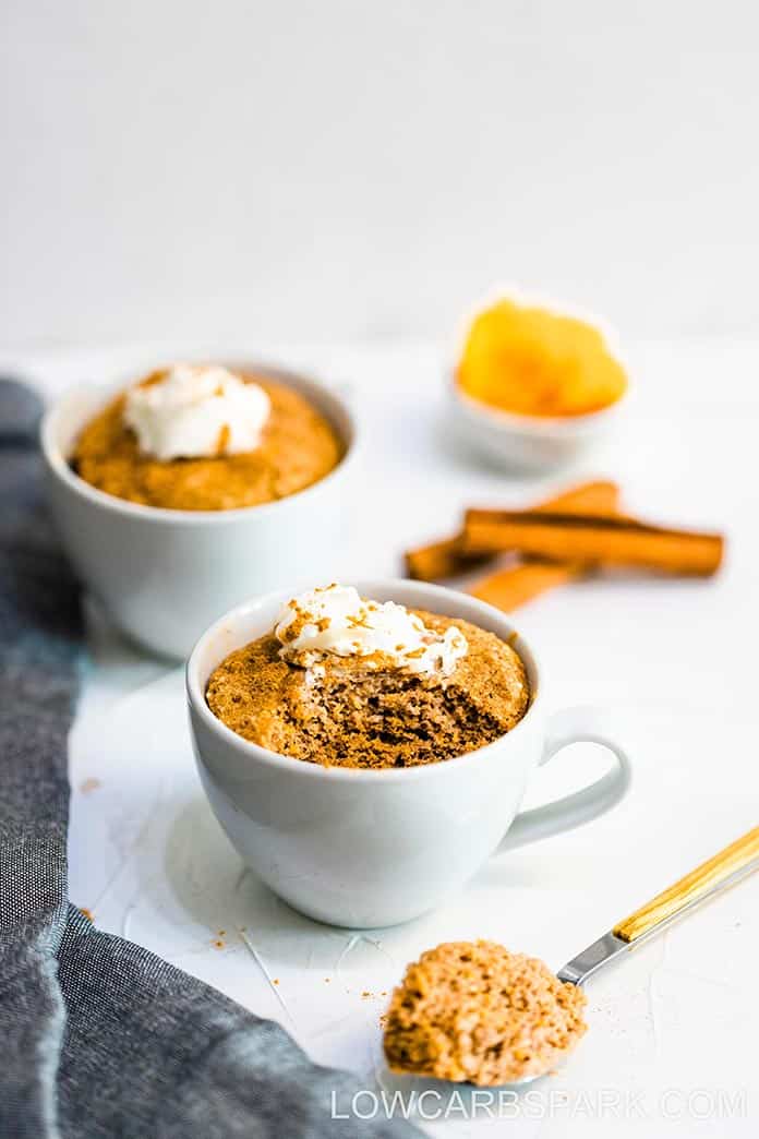 two white teacups filled with orange cake topped with whipped cream and cinnamon on a white table next to cinnamon sticks and a bowl of orange pumpkin puree