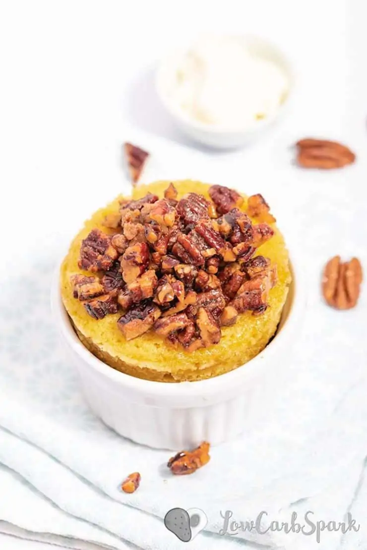 White ramekin with yellow cake inside topped with chopped pecans on a white surface with pecans next to it