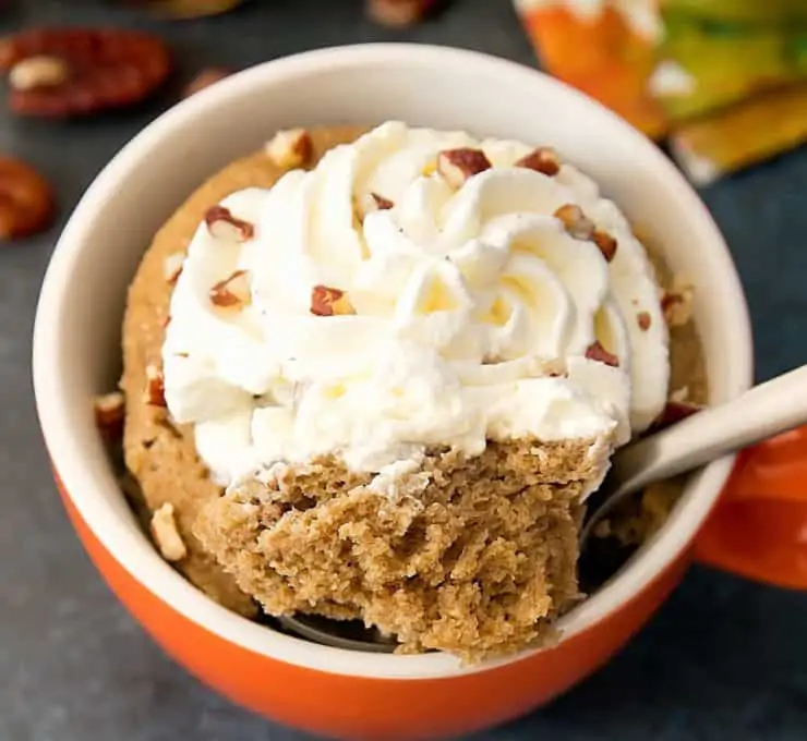 Orange and white ramekin filled with brown cake topped with a spoon takin a bite out with swirled whipped cream with chopped pecans on top