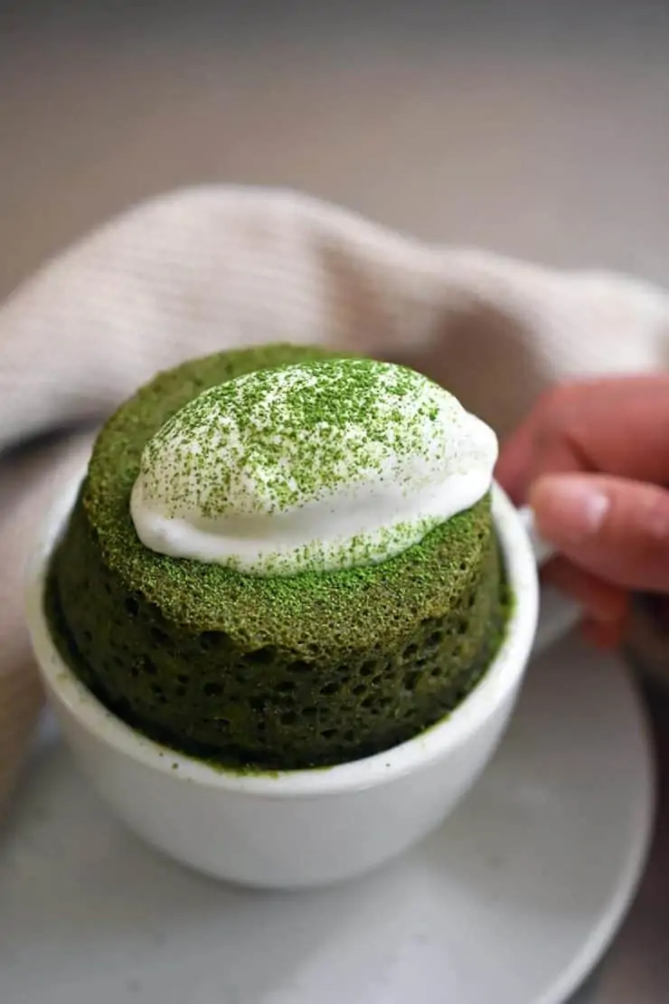 Hand holding a small white tea cup with green matcha cake inside with a dollop of white whipped cream on top sprinkled with green matcha powder next to a white dish towel