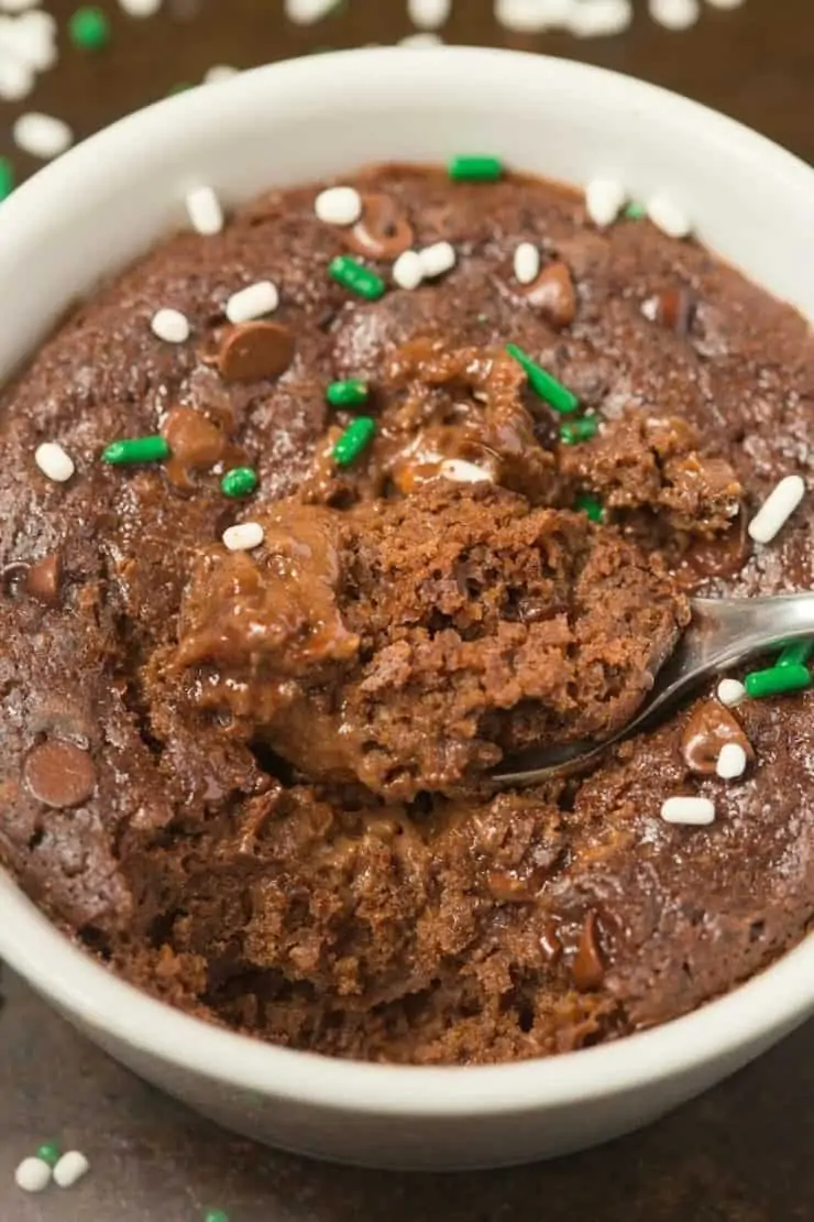 Close up shot of chocolate cake in a white mug with green and white sprinkles on top with a spoon taking a scoop out