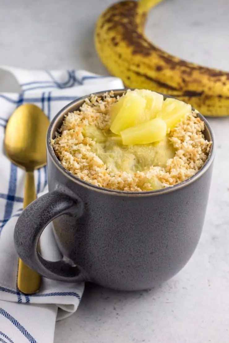 Grey mug with cake inside topped with coconut and pineapple chunks with a gold spoon and blue and white plaid dish towel and banana next to it