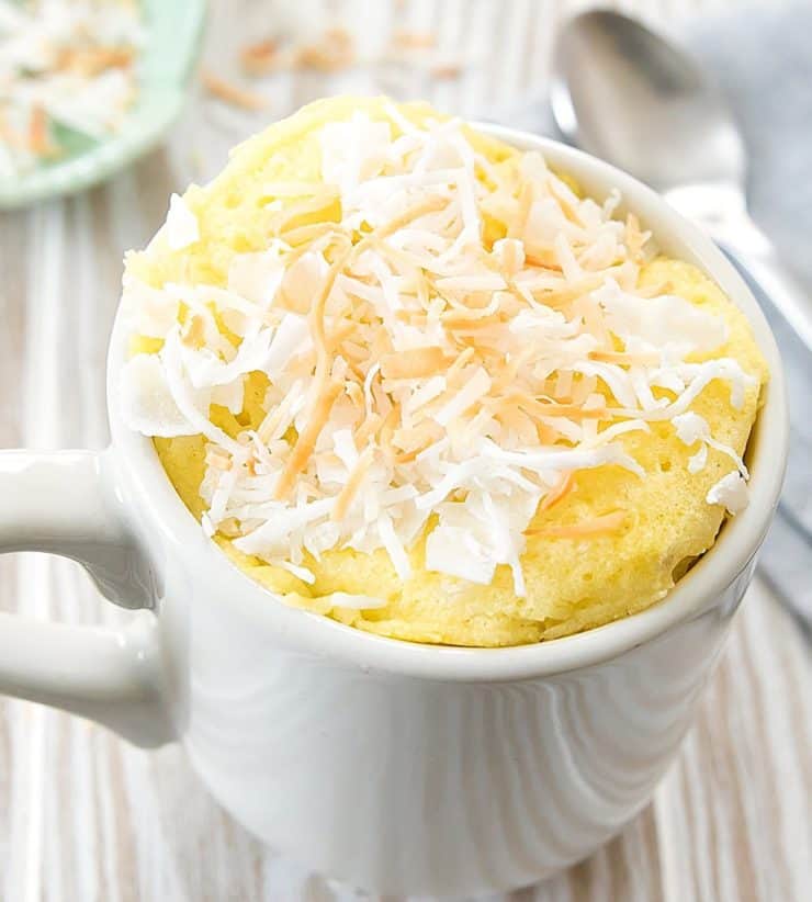 White mug filled with yellow cake topped with shredded white and golden brown coconut on a light wooden surface next to a spoon