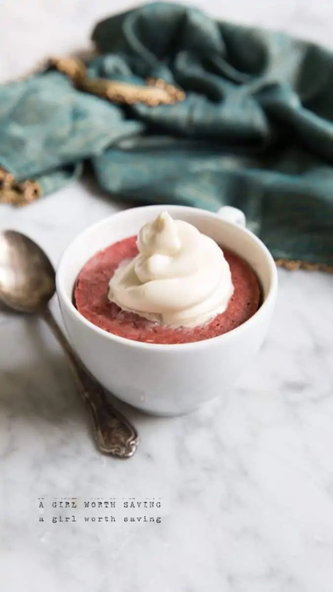 White mug filled with red velvet cake topped with swirled white whipped cream on a marble surface next to a spoon