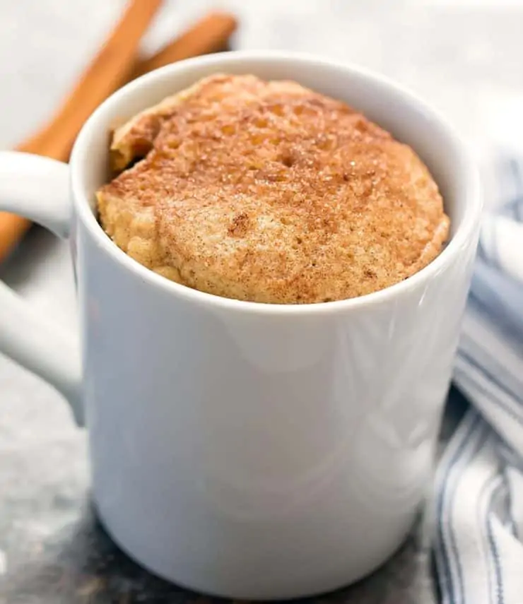 White mug filled with yellow cake topped with cinnamon on a grey surface next to a blue and white striped dish towel next to cinnamon sticks