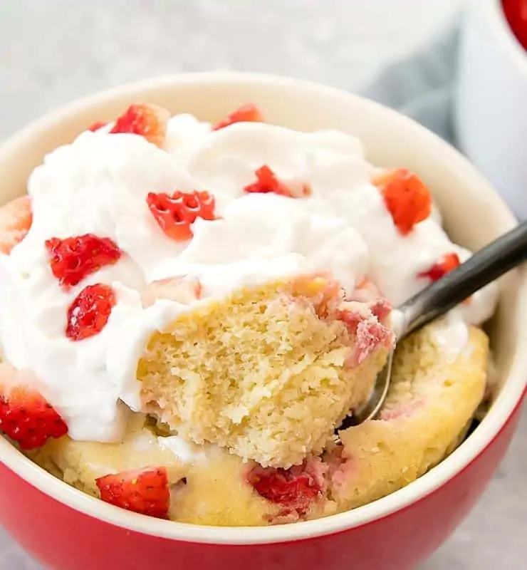 Close up of red and white ramekin filled with yellow cake topped with whipped cream and chopped strawberries with a spoon taking a scoop out of the cake