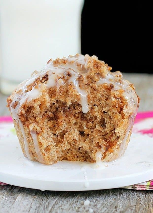 Close up of a bite out of a coffee cake muffin drizzled with white creamy frosting on a white plate with a pink and white dish towel in the background