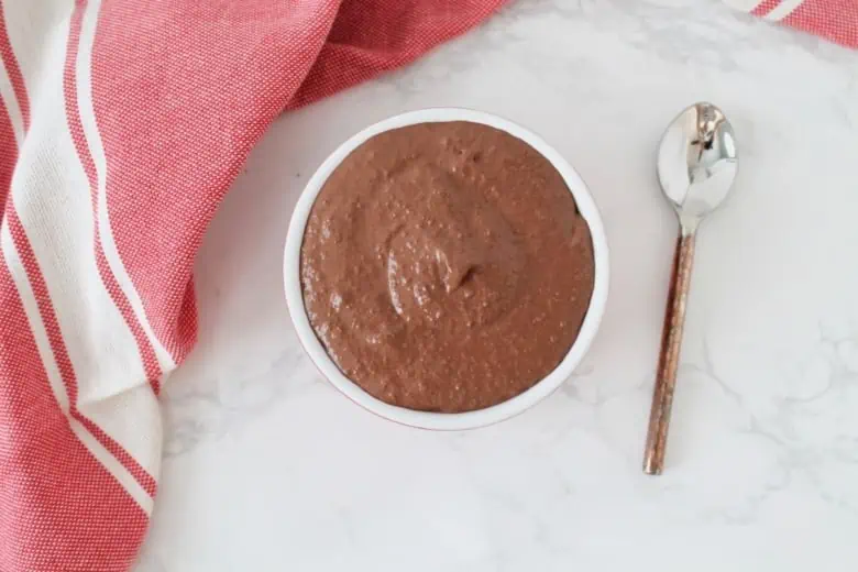 Overhead shot of red ramekin filled with chocolate chia pudding on white marble surface next to spoon and red dish towel