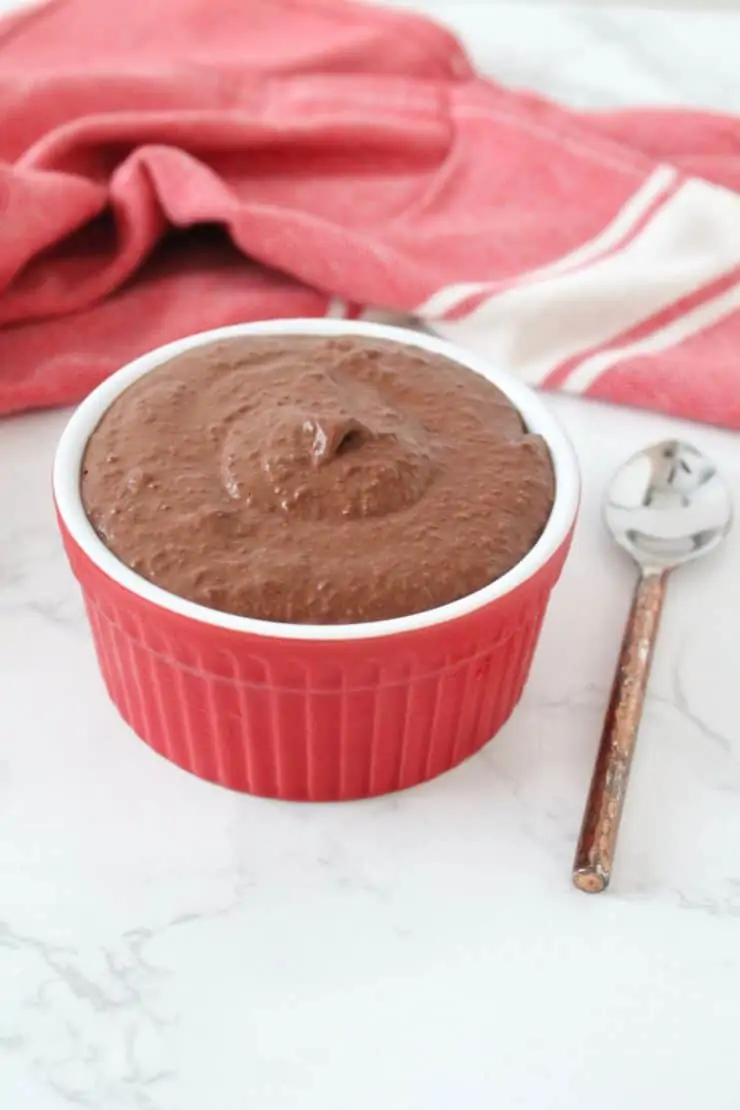 Red ramekin filled with chocolate chia keto pudding on white marble surface next to spoon and red dish towel