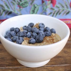 White bowl filled with paleo oats topped with blueberries and cinnamon on a wooden surface next to a spoon with a floral cloth in the background