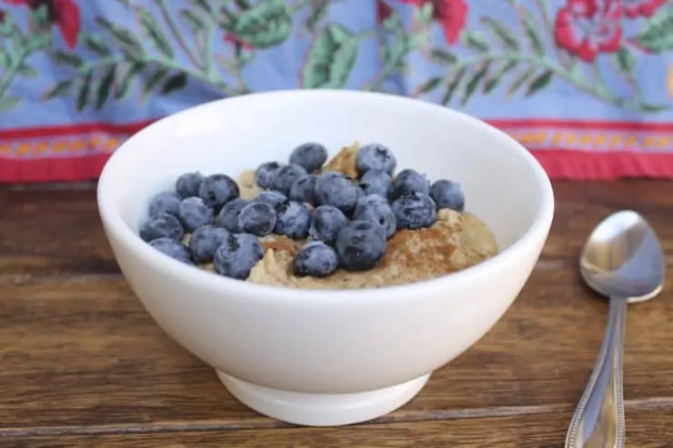 White bowl filled with paleo oats topped with blueberries and cinnamon on a wooden surface next to a spoon with a floral cloth in the background
