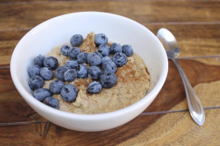 Close up of a white bowl filled with paleo oats topped with blueberries and cinnamon on a wooden surface next to a spoon