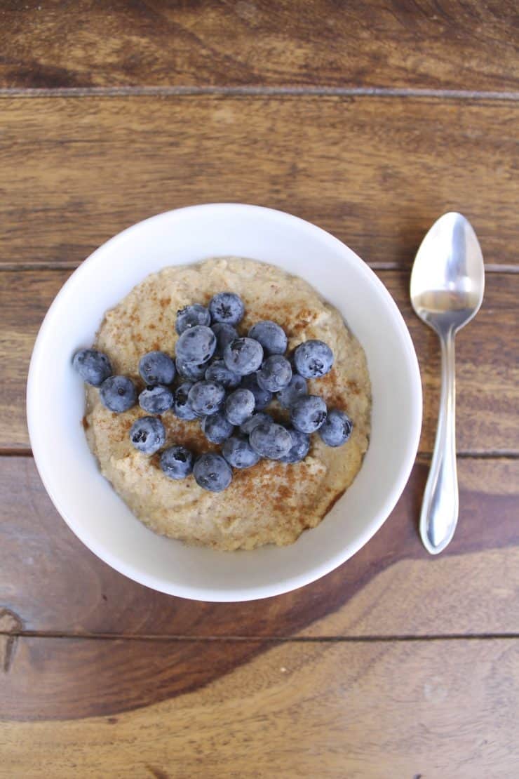 Overhead shot of a white bowl filled with paleo oatmeal topped with blueberries and cinnamon on a wooden surface next to a spoon