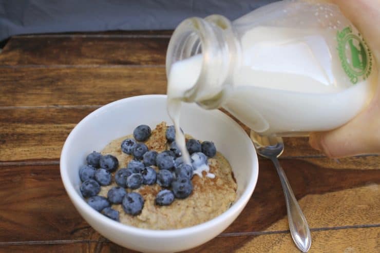 White bowl filled with paleo oatmeal topped with blueberries and cinnamon on a wooden surface with a hand pouring milk onto the oats out of a glass milk jug