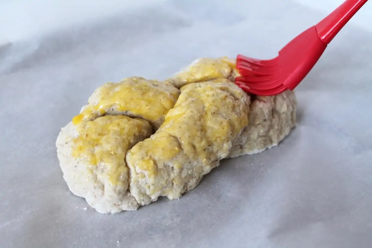 Hand brushing egg yolk with a red silicone brush onto an uncooked loaf of braided challah