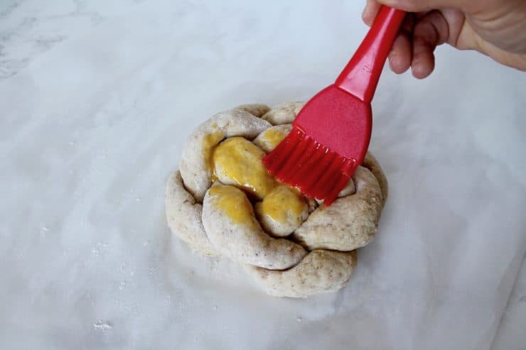 Hand brushing egg yolk with a red silicone brush onto an uncooked loaf of round braided challah