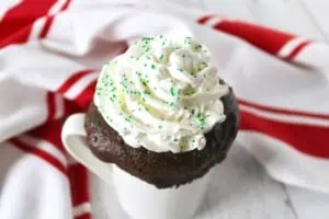 Close up of peppermint mocha keto mug cake with whipped cream on top sprinkled with green sprinkles in a white mug on a white wooden surface with a red and white striped dish towel in the background