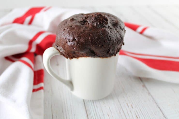 Close up of peppermint mocha keto mug cake in a white mug on a white wooden surface with a red and white striped dish towel in the background