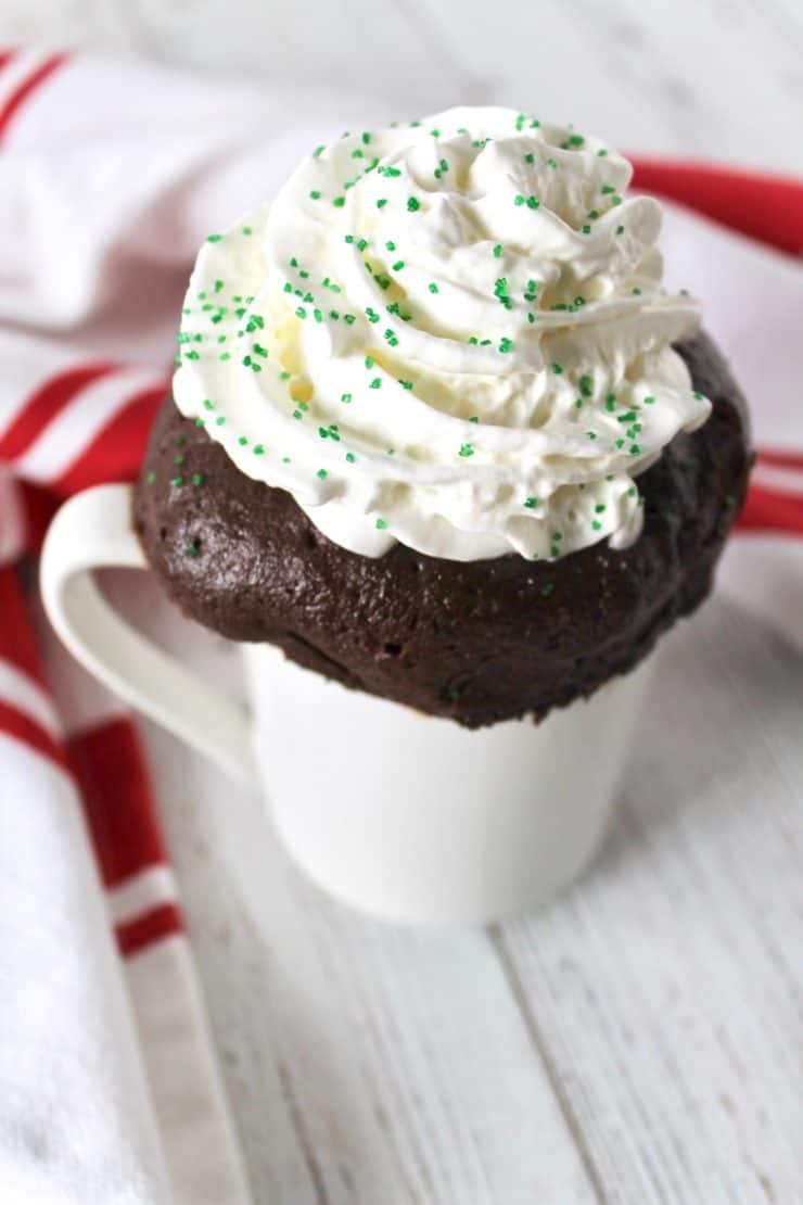 Close up of peppermint mocha keto mug cake with whipped cream on top sprinkled with green sprinkles in a white mug on a white wooden surface with a red and white striped dish towel in the background