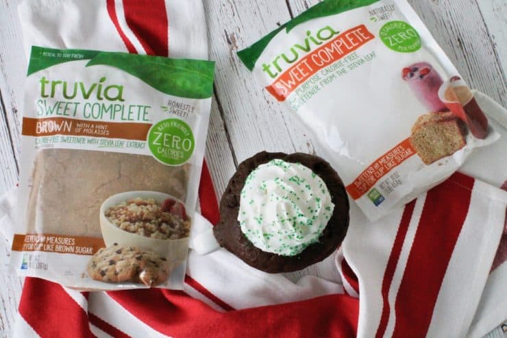 Overhead shot of peppermint mocha mug cake with whipped cream and green sprinkles on top next to two bags of truvia sweet complete sweetener on a white wooden surface next to a red and white striped towel