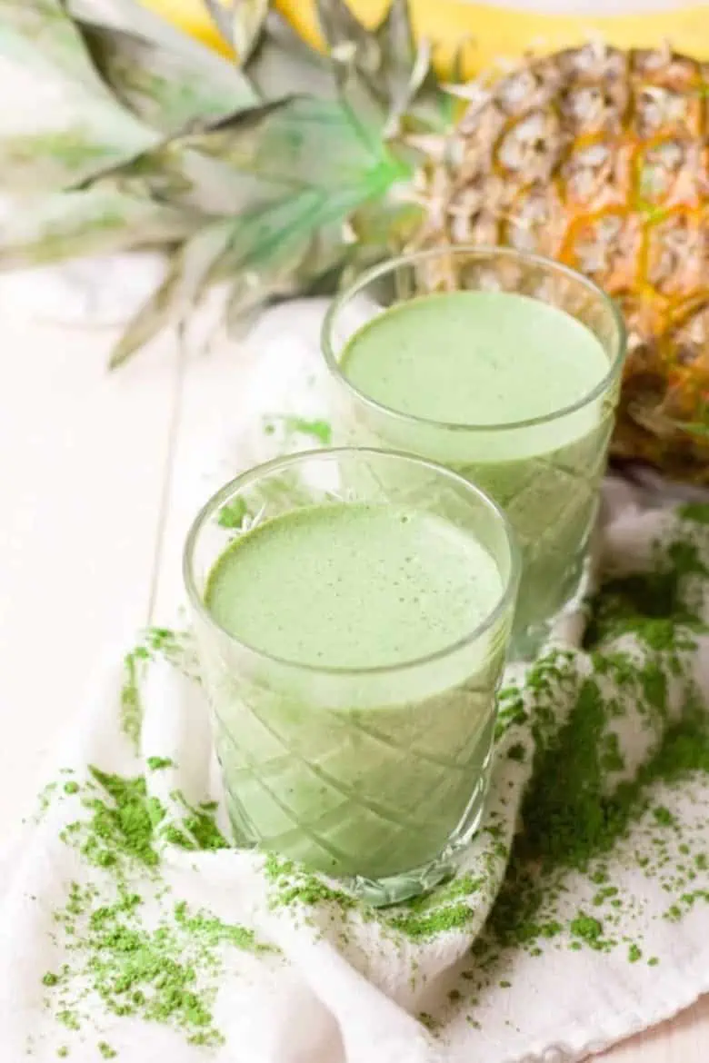 two glasses with green moringa smoothies in front of a pineapple and banana