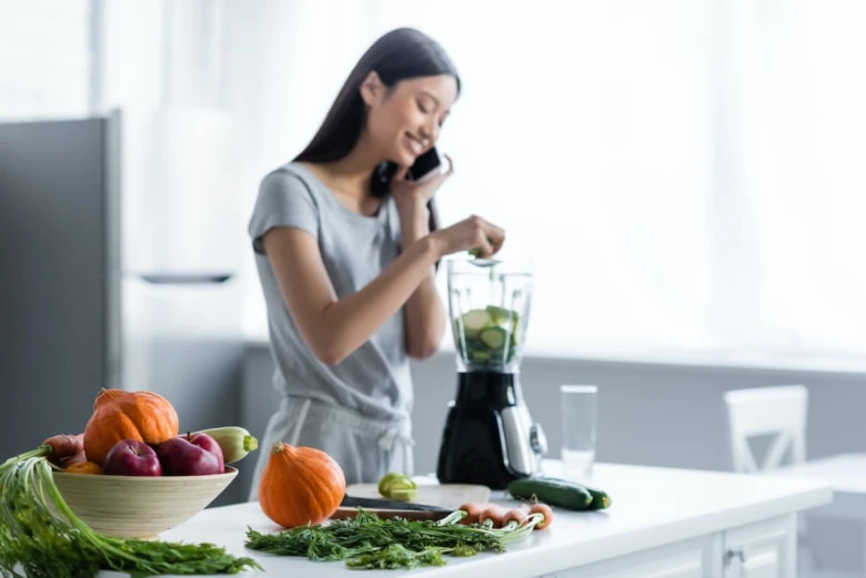 A woman talking on a cell phone while placing cucumbers in a blender