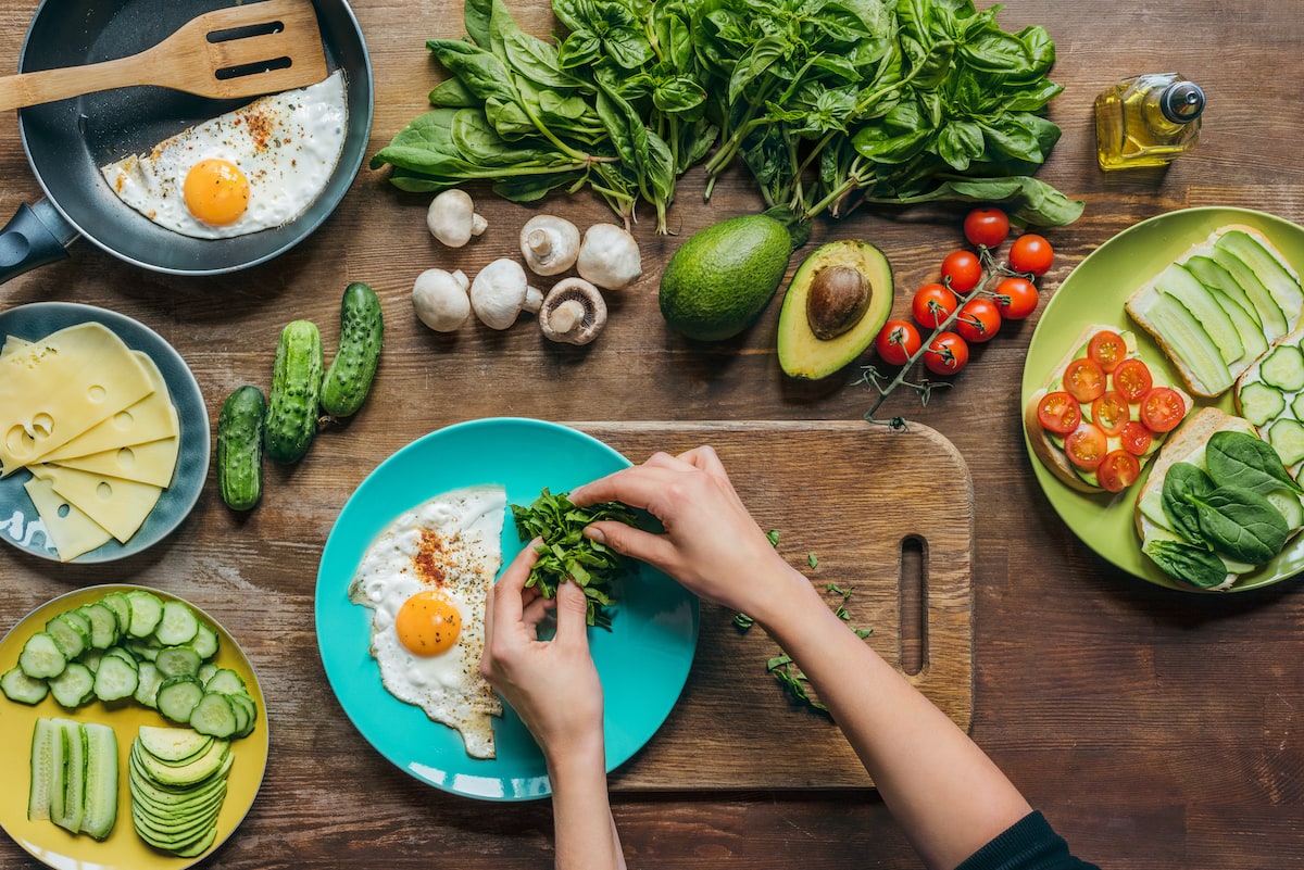 overhead shot of various vegetables on a wooden table, a teal plate with a fried egg on it with a woman's hands placing greens on the plate