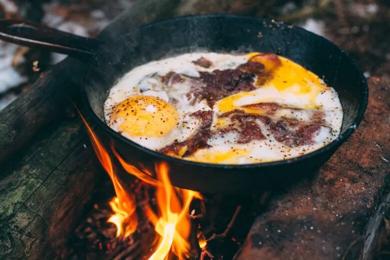 Cast iron skillet with eggs and bacon in it over an open outdoor fire