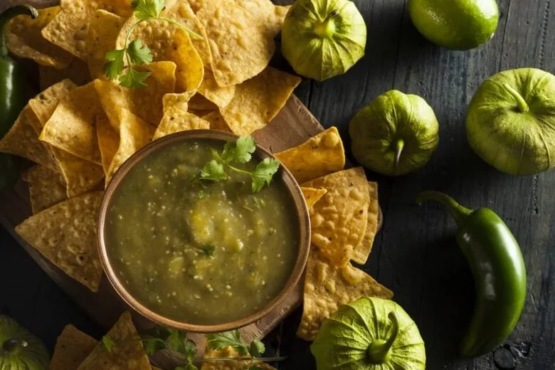 Overheat shot of tomatillo salsa surrounded by chips next to raw tomatillos