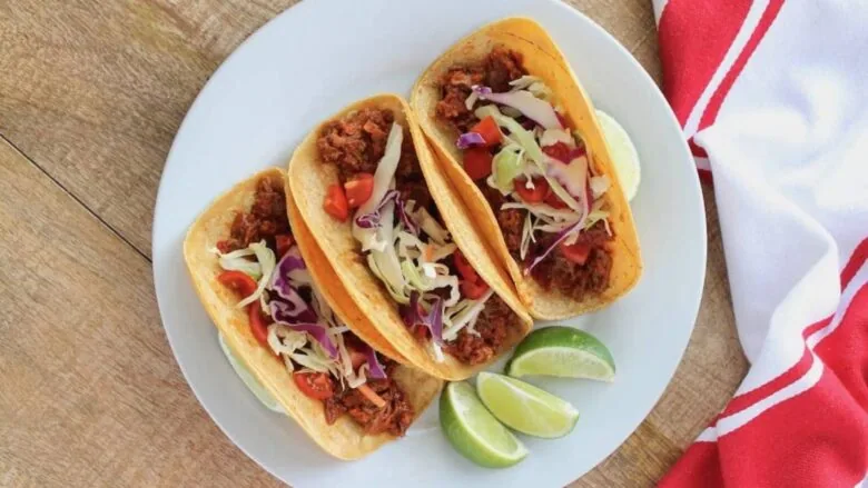 Three tacos on a white plate with lime wedges.