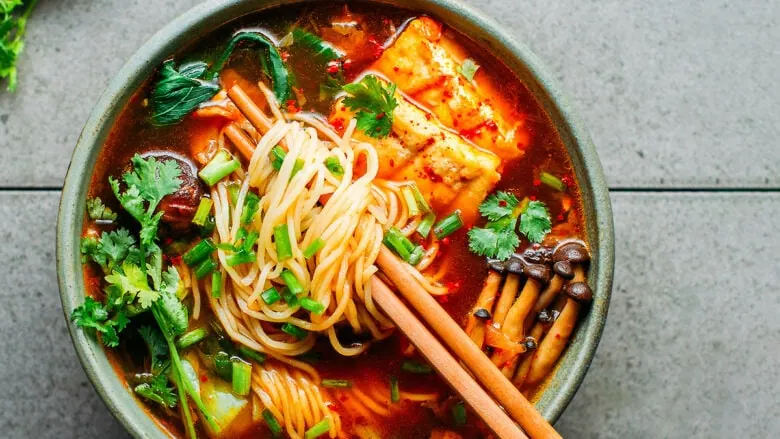 A bowl of Asian noodle soup with chopsticks and kimchi.