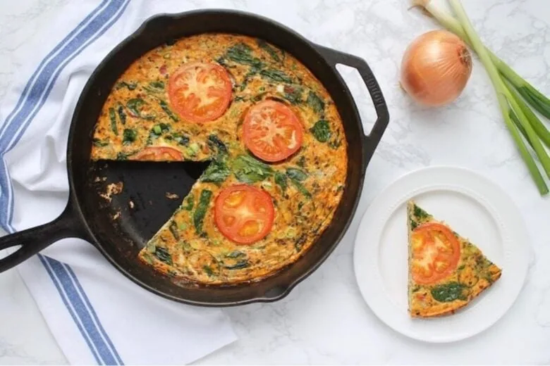 A spinach and tomato frittata in a cast iron skillet, with a slice on a white plate and raw ingredients nearby.