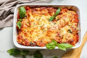 Baked lasagna in a white dish garnished with fresh basil leaves on a light surface with a wooden spoon and cloth napkin.