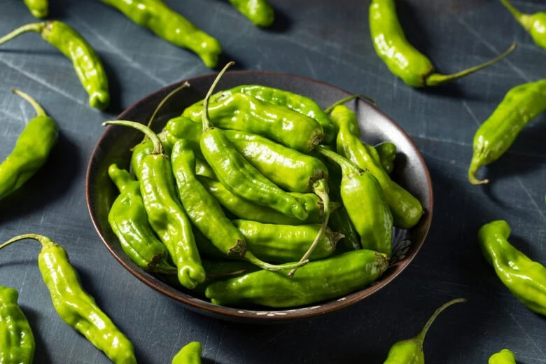 A bowl of fresh green shishito peppers, known for their health benefits, on a dark textured surface.