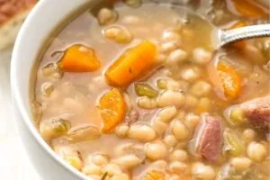 A close-up of a bowl of bean soup containing carrots, celery, and chunks of meat, with a spoon partially visible in the soup.