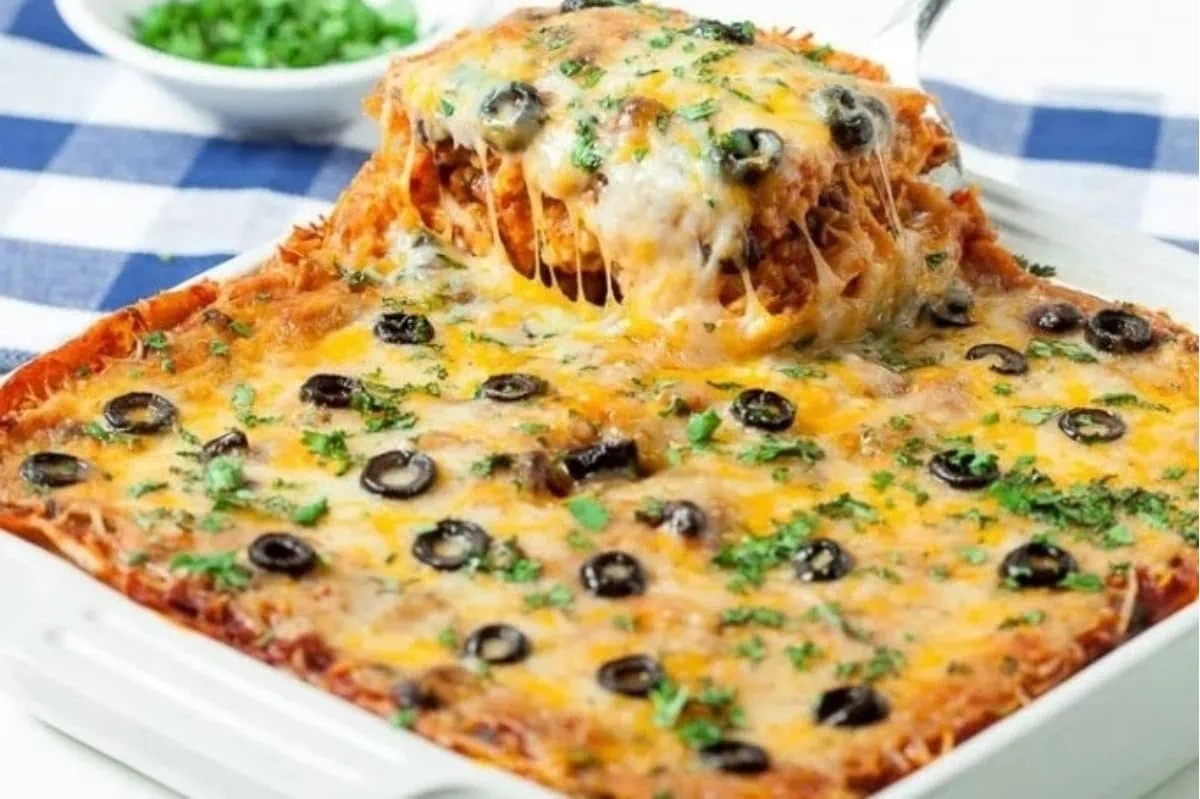 A baked casserole topped with melted cheese, black olives, and herbs, being served in a square ceramic dish with a portion lifted out, placed on a checkered blue and white tablecloth.