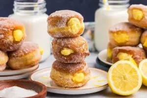 A stack of three sugar-coated, lemon cream-filled doughnuts on a plate, with more doughnuts and lemon halves around, and glass jars of milk in the background.