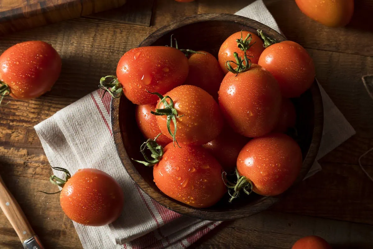 A wooden bowl filled with ripe Roma tomatoes, known for their numerous health benefits, is placed on a cloth on a wooden table. Additional tomatoes and a knife are scattered around.