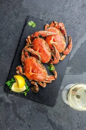 Three cooked crabs served on a black slate with lemon wedges and parsley, accompanied by a glass of white wine.