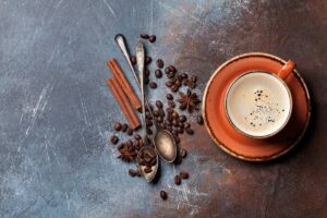 A cup of coffee sits on a saucer with coffee beans, cinnamon sticks, star anise, and two spoons on a textured surface.